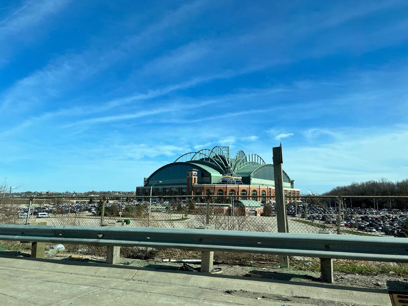 American Family Field seen from the highway, its arched roof against a clear blue sky above the parking lots