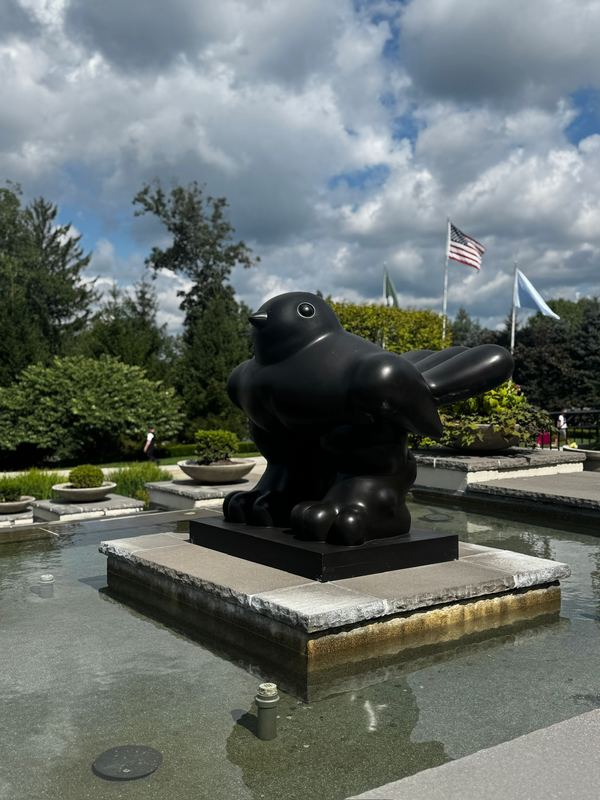 A rounded bronze bird sculpture on a plinth in a fountain pool, flags and clouds behind