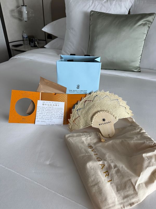 A Bvlgari gift laid out on a white bed: an orange box, a handwritten card, a woven straw fan and a dust bag, a Ritz-Carlton Yacht Collection bag behind