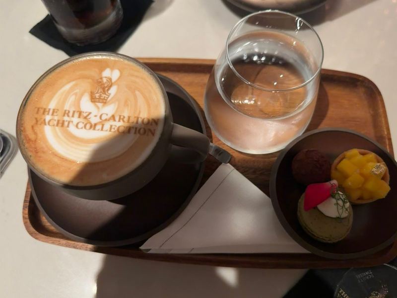 A cappuccino with the Ritz-Carlton Yacht Collection crest in the foam, two small pastries on a tray beside it