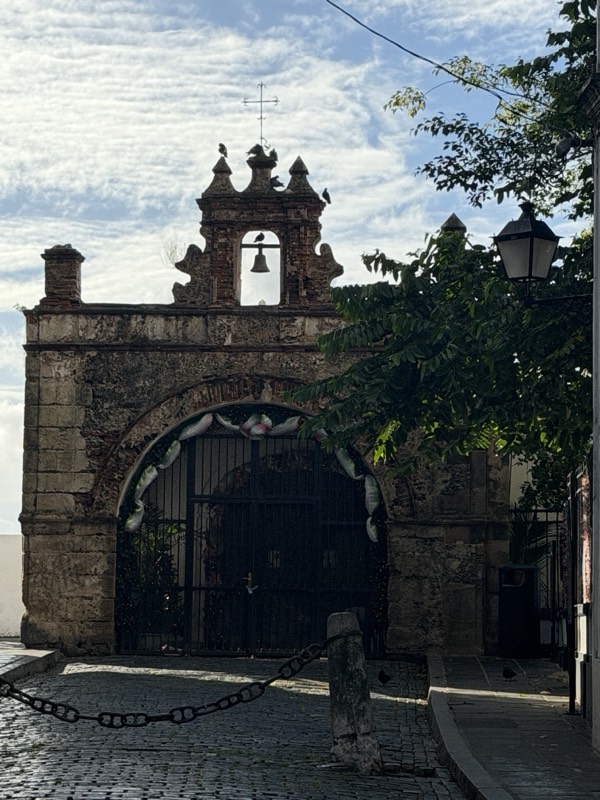 The Capilla del Santo Cristo de la Salud: a stone bell-gable above an iron gate at the foot of Calle del Cristo