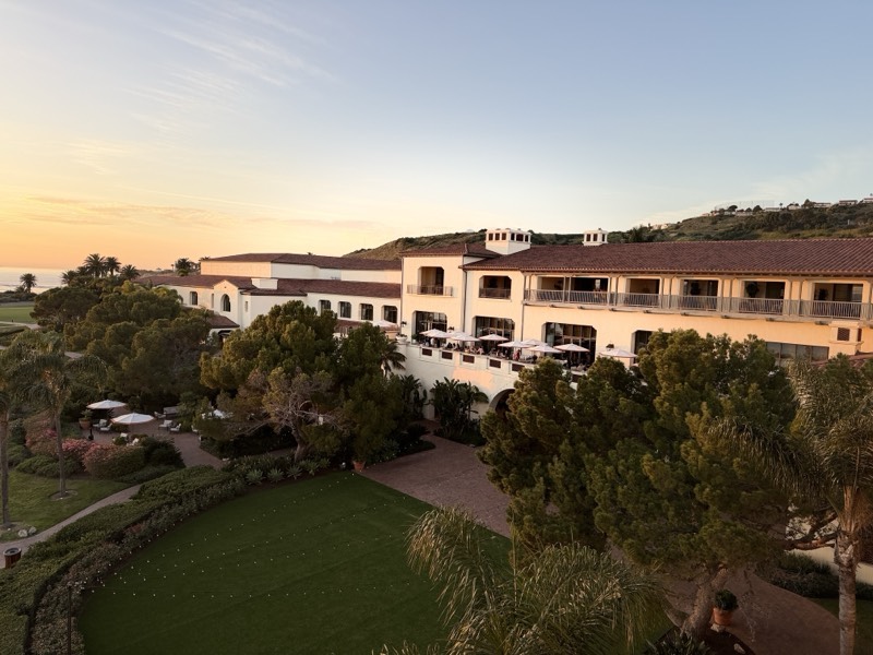 A clifftop resort at dusk above the ocean, terrace umbrellas and lawn below a tiled roofline