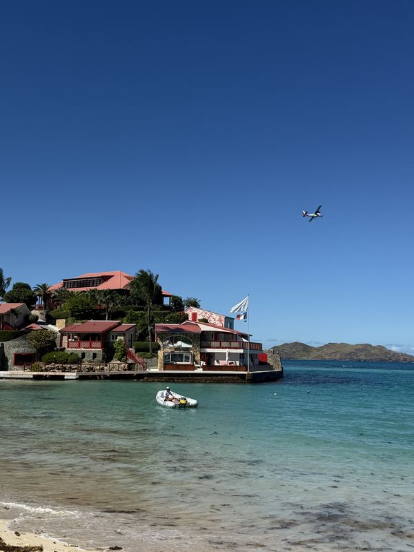 Red-roofed hotel on a rocky point in a turquoise bay, a plane on approach overhead, St. Jean Bay, St. Barts