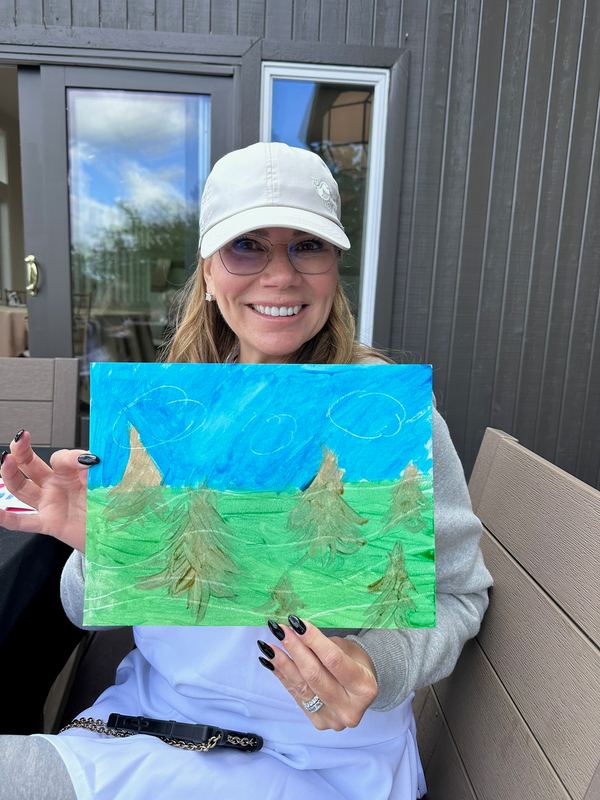 Jeannie in a white cap holding up her painting of pine trees under a blue sky