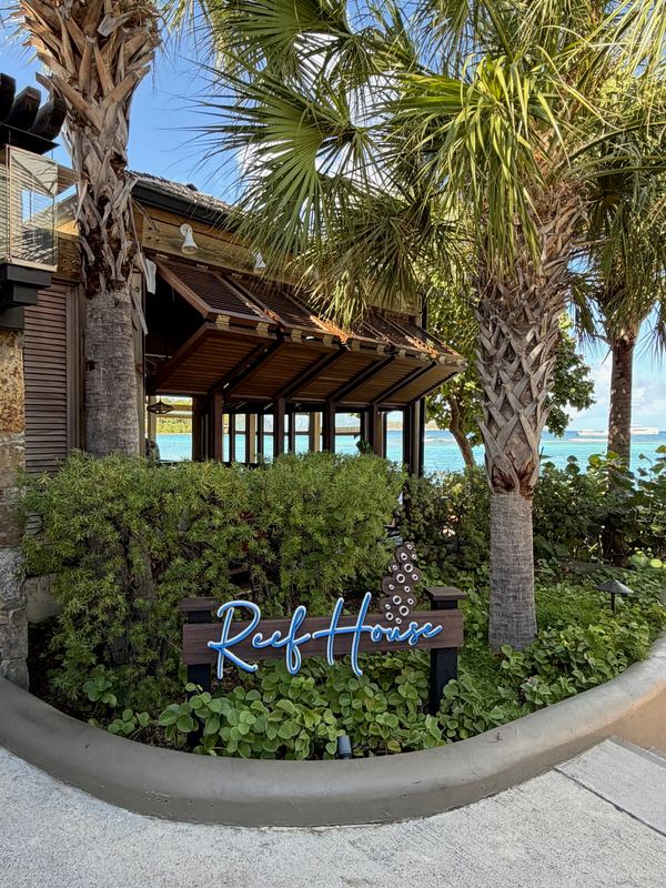 The Reef House sign under palms at Rosewood Little Dix Bay, the sea beyond