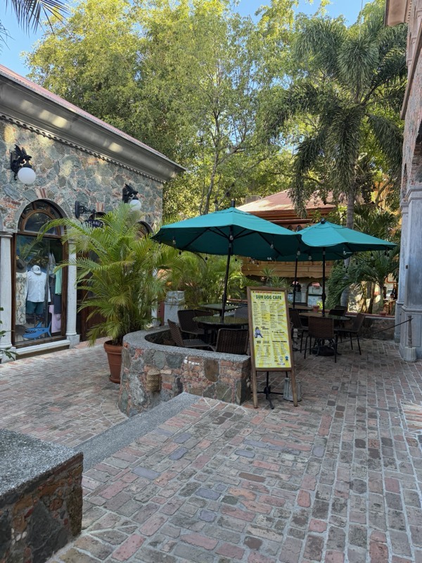 A stone-fronted shop courtyard in Cruz Bay with teal umbrellas, brick paving and palms