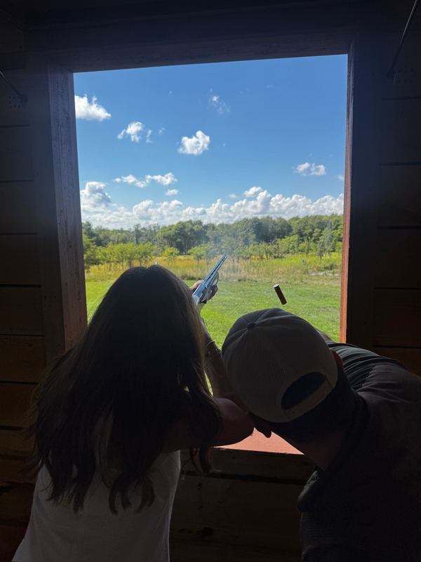 Jeannie firing a shotgun from the window of a wooden shooting stand, smoke in the air, the instructor beside her