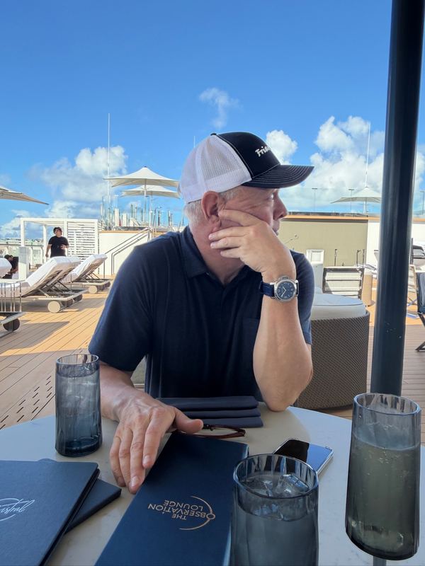 Steve in a cap at a table on deck in the sun, glasses of water and menus in front of him