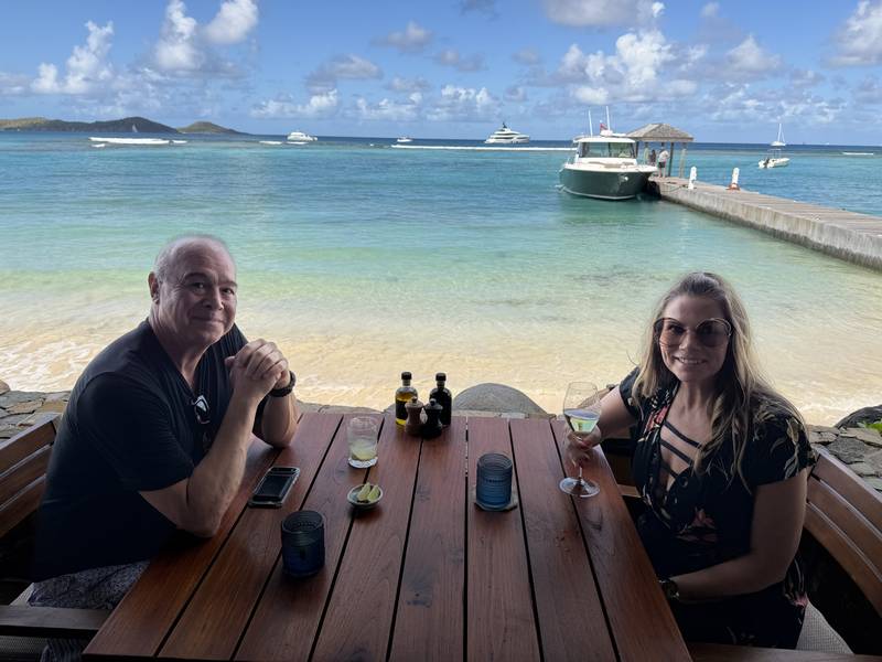 Steve and Jeannie at a wooden table on the sand at the Reef House, the tender at the dock and yachts on the water behind