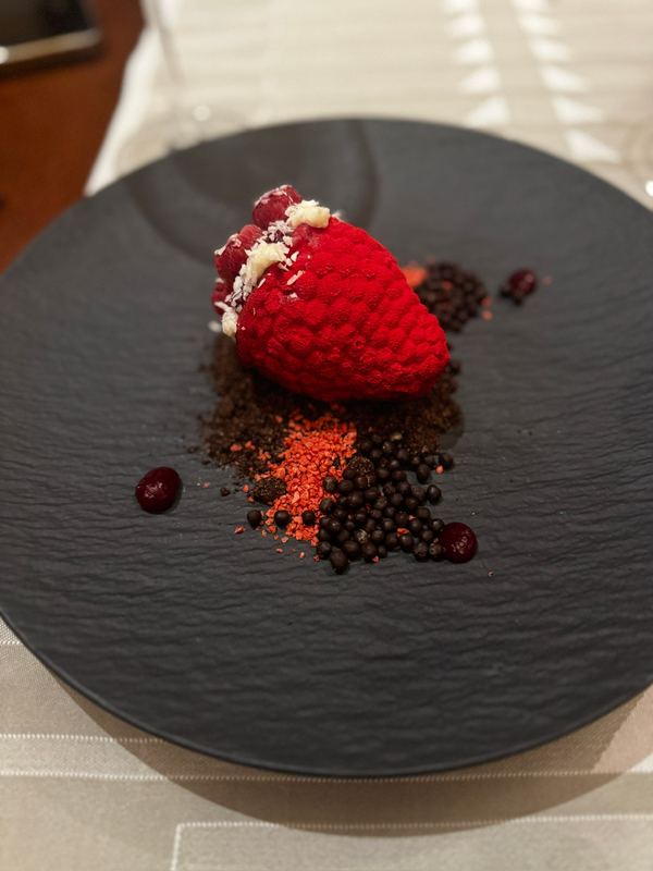 A red strawberry-shaped dessert on a black slate plate with crumbs and berries