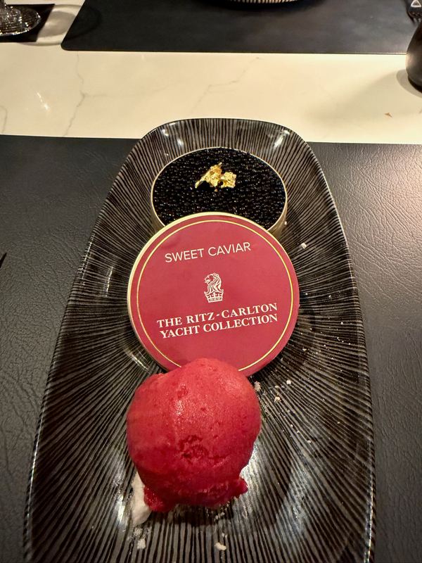 A dessert plated as sweet caviar: dark pearls under a red Ritz-Carlton Yacht Collection tin lid, with a scoop of red sorbet