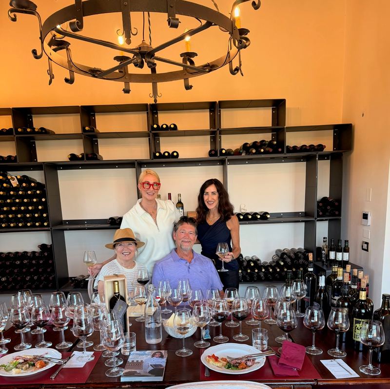 Four at the tasting table in the cellar, bottle racks behind and a row of glasses in front