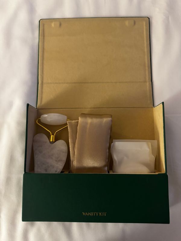 An open green vanity kit box on the bed holding a jade roller, a gua sha and pouches