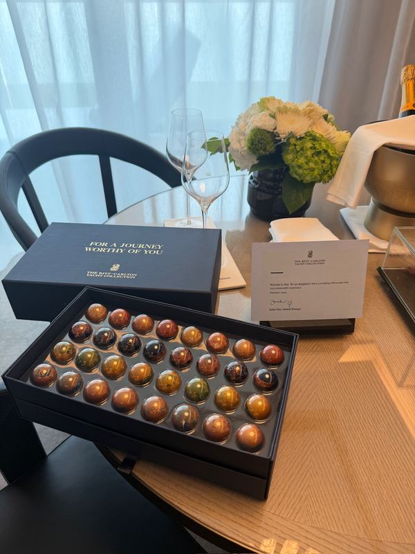 An open box of glossy chocolates on a table in the suite, a card and a small vase of flowers behind